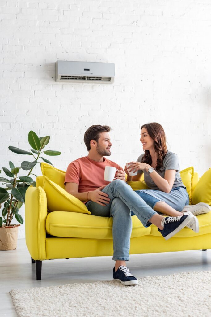 handsome man with pretty girlfriend holding cups while sitting on sofa under air conditioner at home